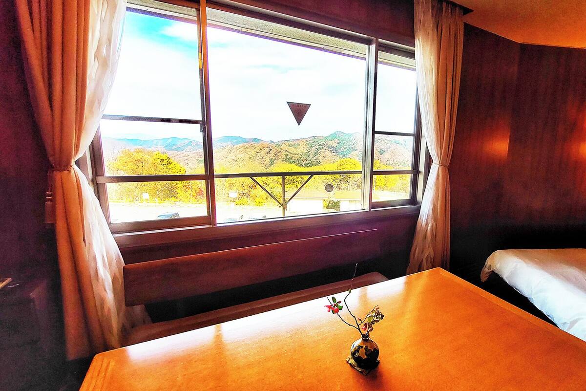Habitación de hotel con cama, una mesita con un jarrón, ventana con cortinas y vista a un paisaje montañoso.
