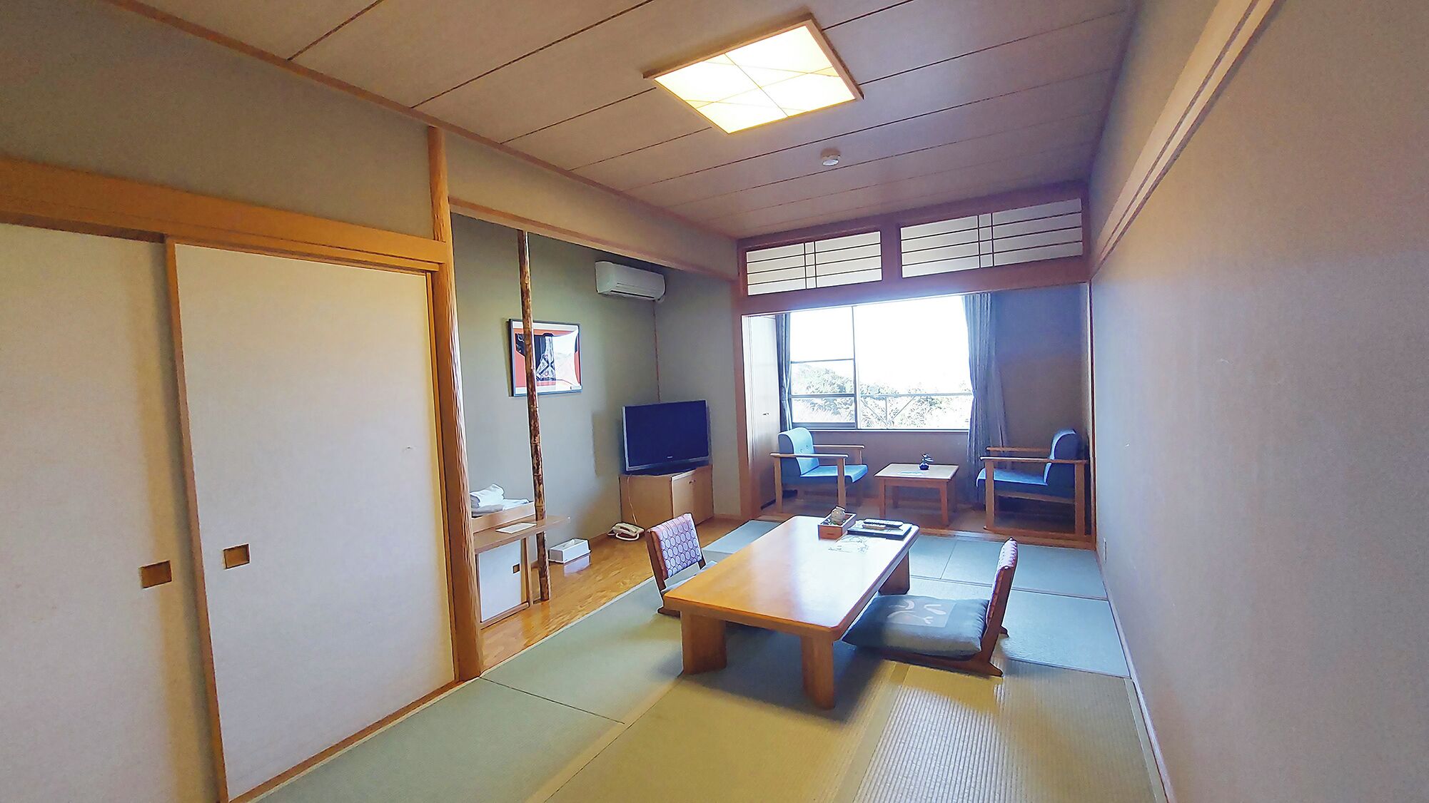 A traditional Japanese room with tatami flooring, a low table, chairs, a television, and a sliding door.