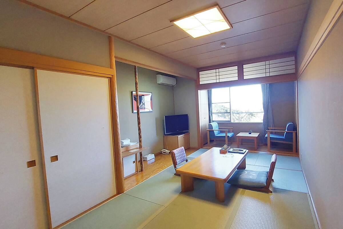 Una habitación japonesa tradicional con suelo de tatami, una mesa baja, sillas, un televisor y una puerta corredera.