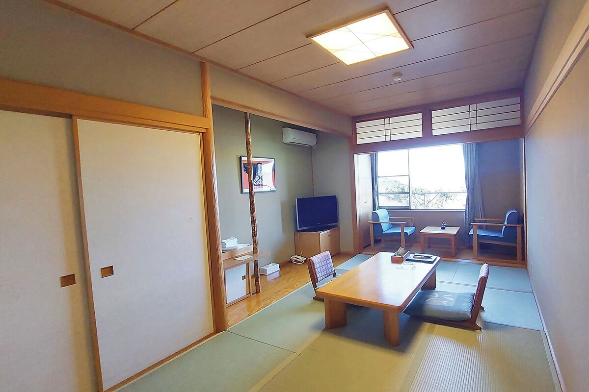 Une pièce de style japonais traditionnel, avec un sol recouvert de tatamis, une table basse, des chaises, un téléviseur et une porte coulissante.