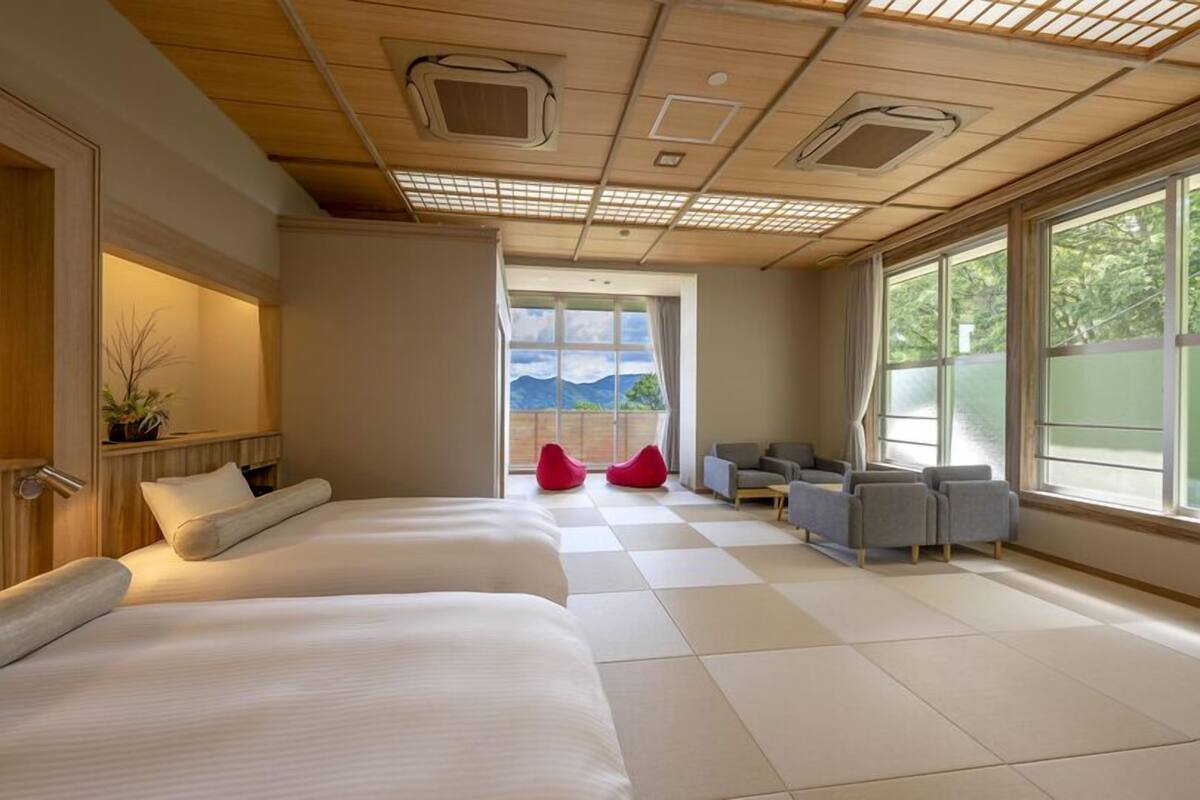 Habitación de hotel con dos camas, una zona de estar con sillas, un ventanal con vistas e iluminación empotrada en el techo.