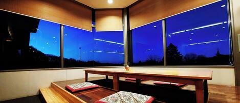 Una habitación moderna con una mesa grande y bancos, ventanas amplias con vistas al cielo nocturno.
