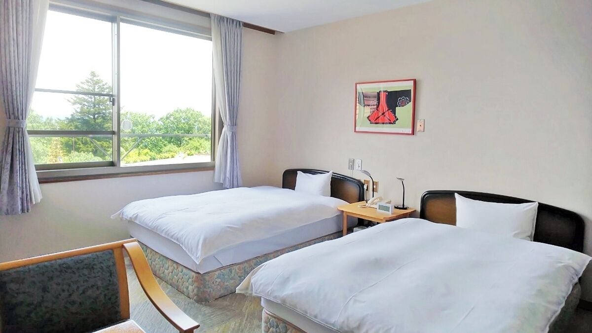 A hotel room with two beds, a bedside table with a lamp, a window with curtains, and a framed picture on the wall.