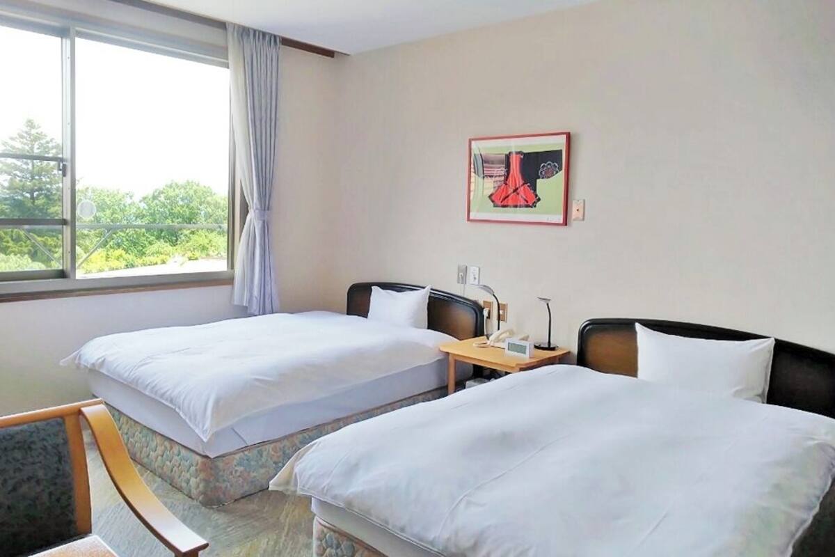 Habitación de hotel con dos camas, una mesita de noche con lámpara, una ventana con cortinas y un cuadro enmarcado en la pared.