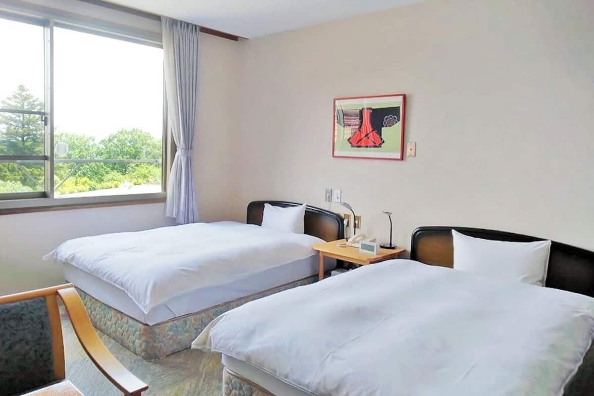 A hotel room with two beds, a bedside table with a lamp, a window with curtains, and a framed picture on the wall.