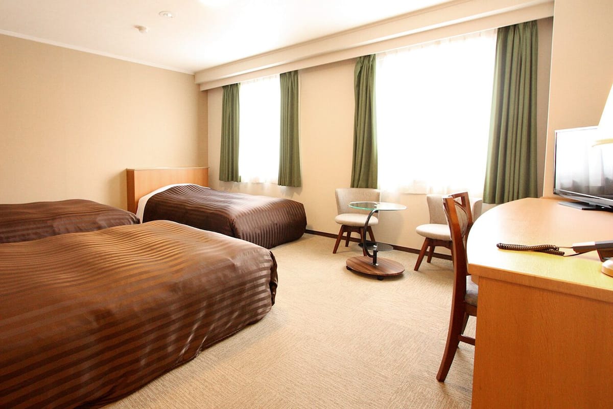 A hotel room with two beds, a desk, a chair, a small table, and a television.