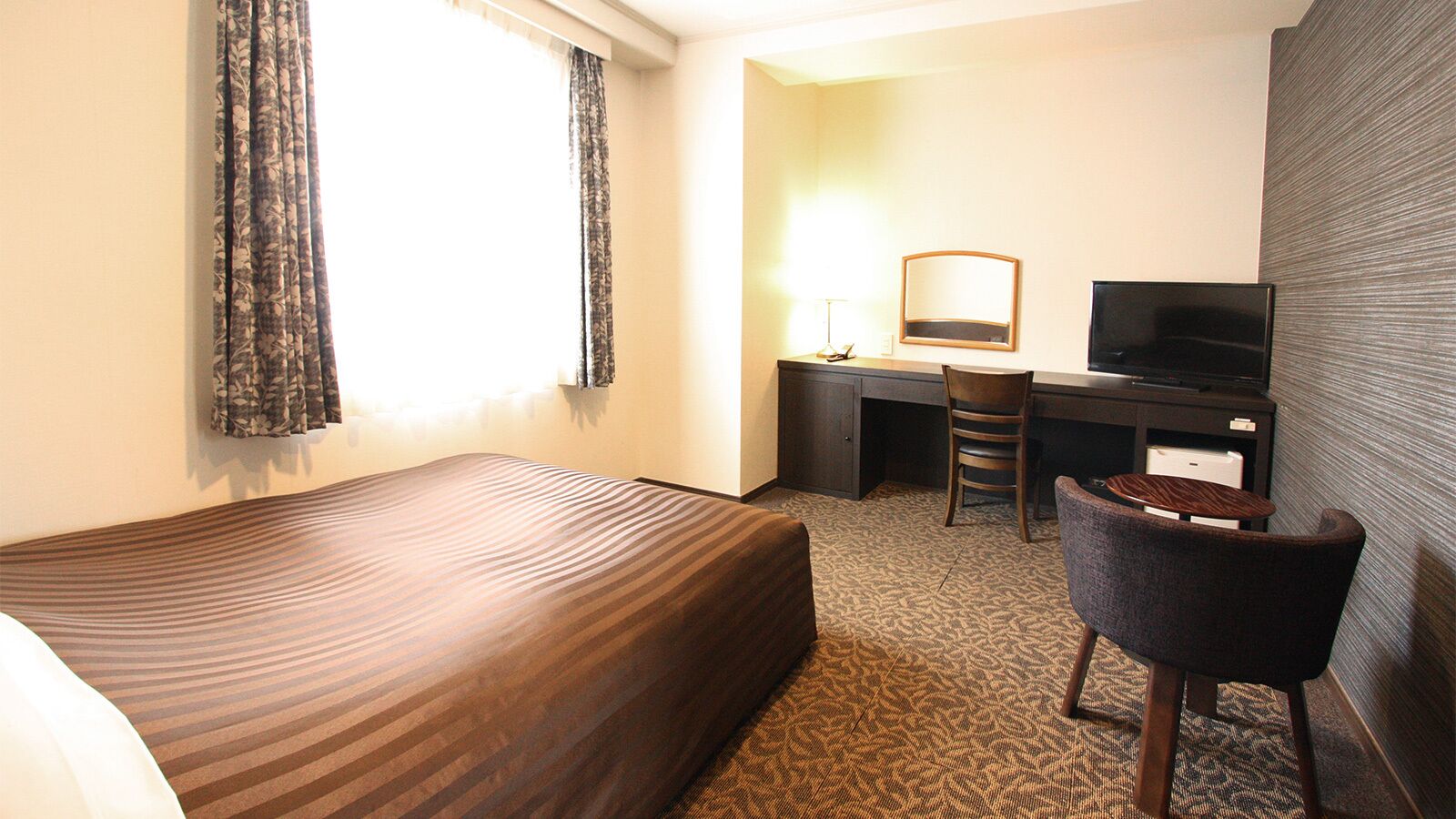 A hotel room with a bed, a desk with a chair, a television, and a window with curtains.