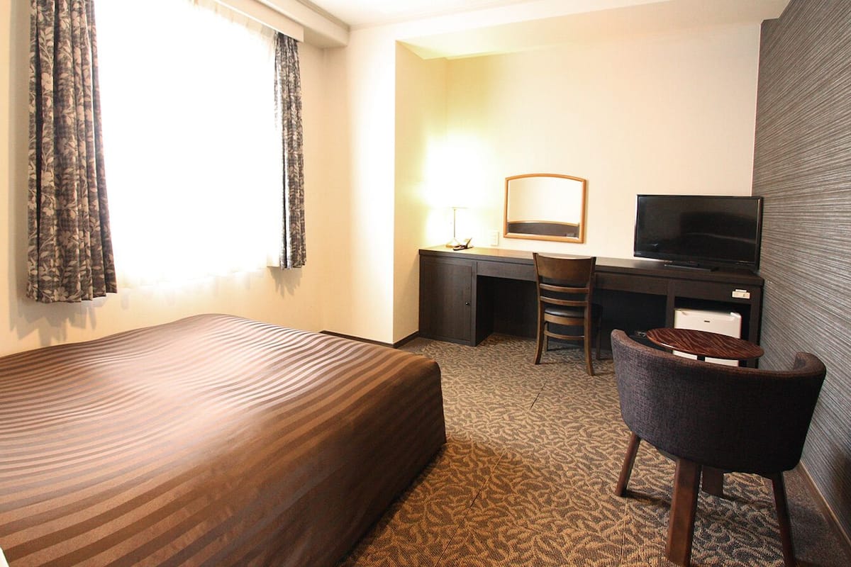 A hotel room with a bed, a desk with a chair, a television, and a window with curtains.