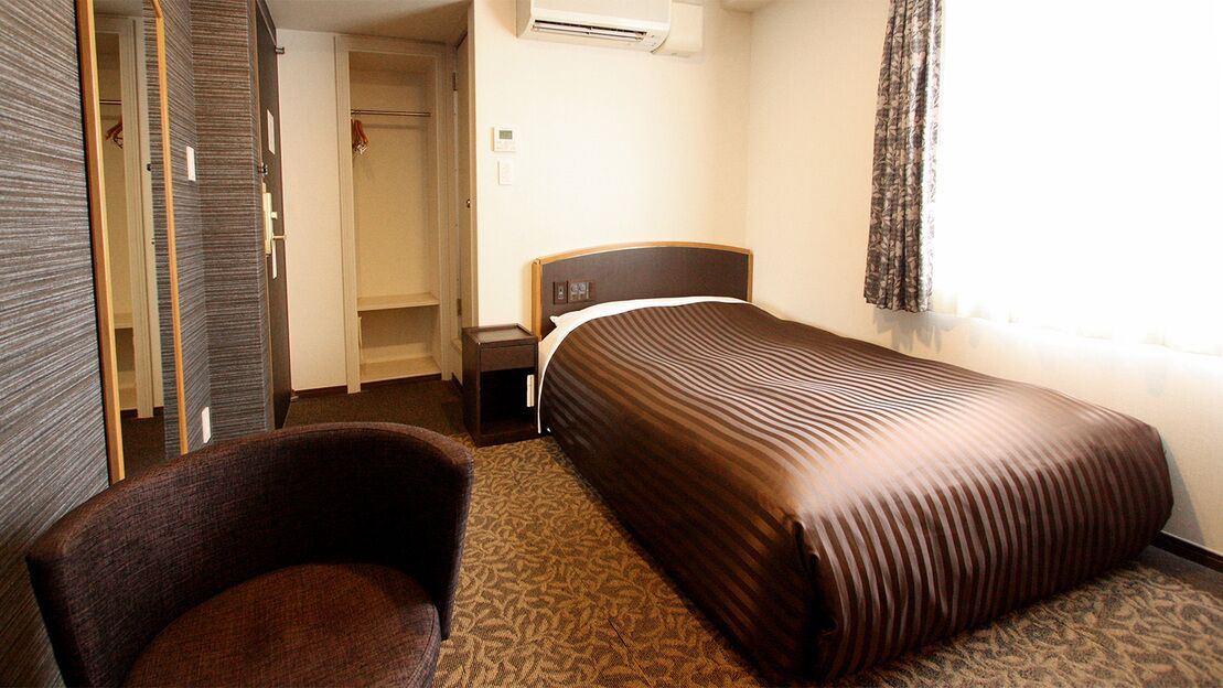 A hotel room with a bed, a chair, a wardrobe, and a nightstand.