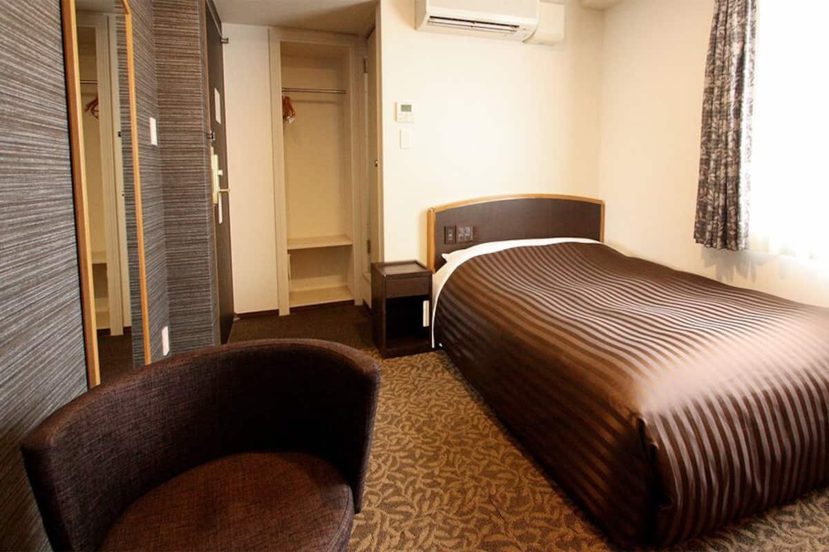 A hotel room with a bed, a chair, a wardrobe, and a nightstand.