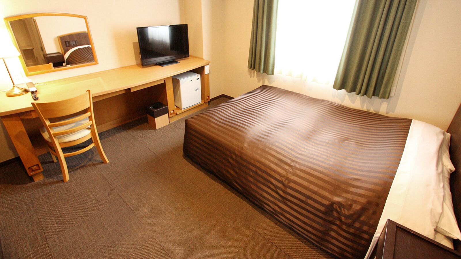 A hotel room with a bed, a desk with a television, a chair, and a small refrigerator.