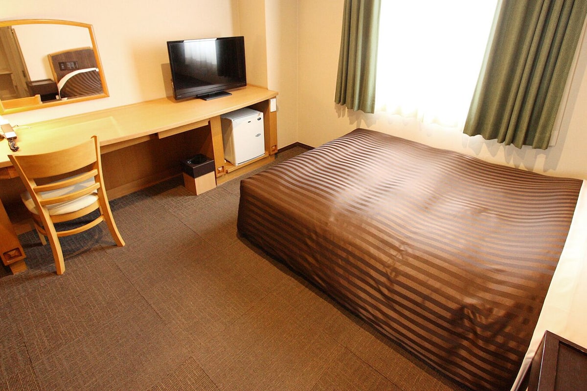 A hotel room with a bed, a desk with a television, a chair, and a small refrigerator.