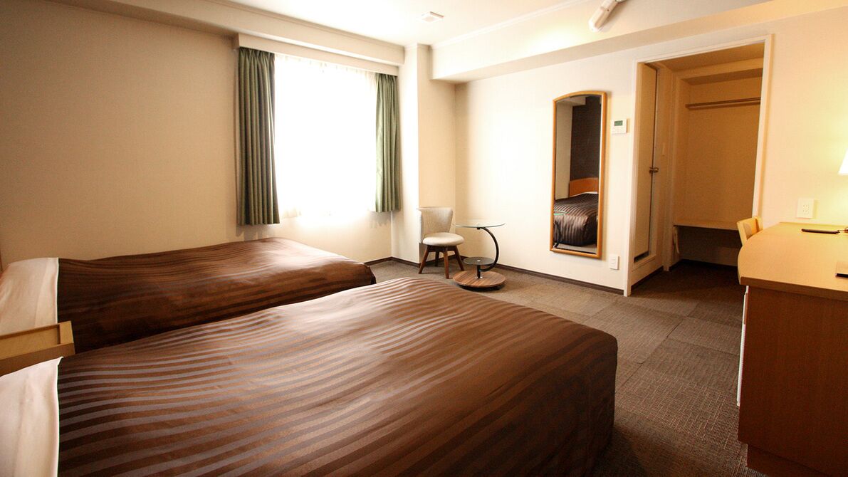 A hotel room with two beds, a chair, a small table, a mirror, and a door leading to a bathroom.