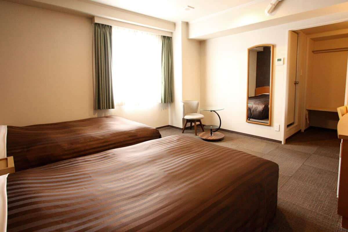 A hotel room with two beds, a chair, a small table, a mirror, and a door leading to a bathroom.