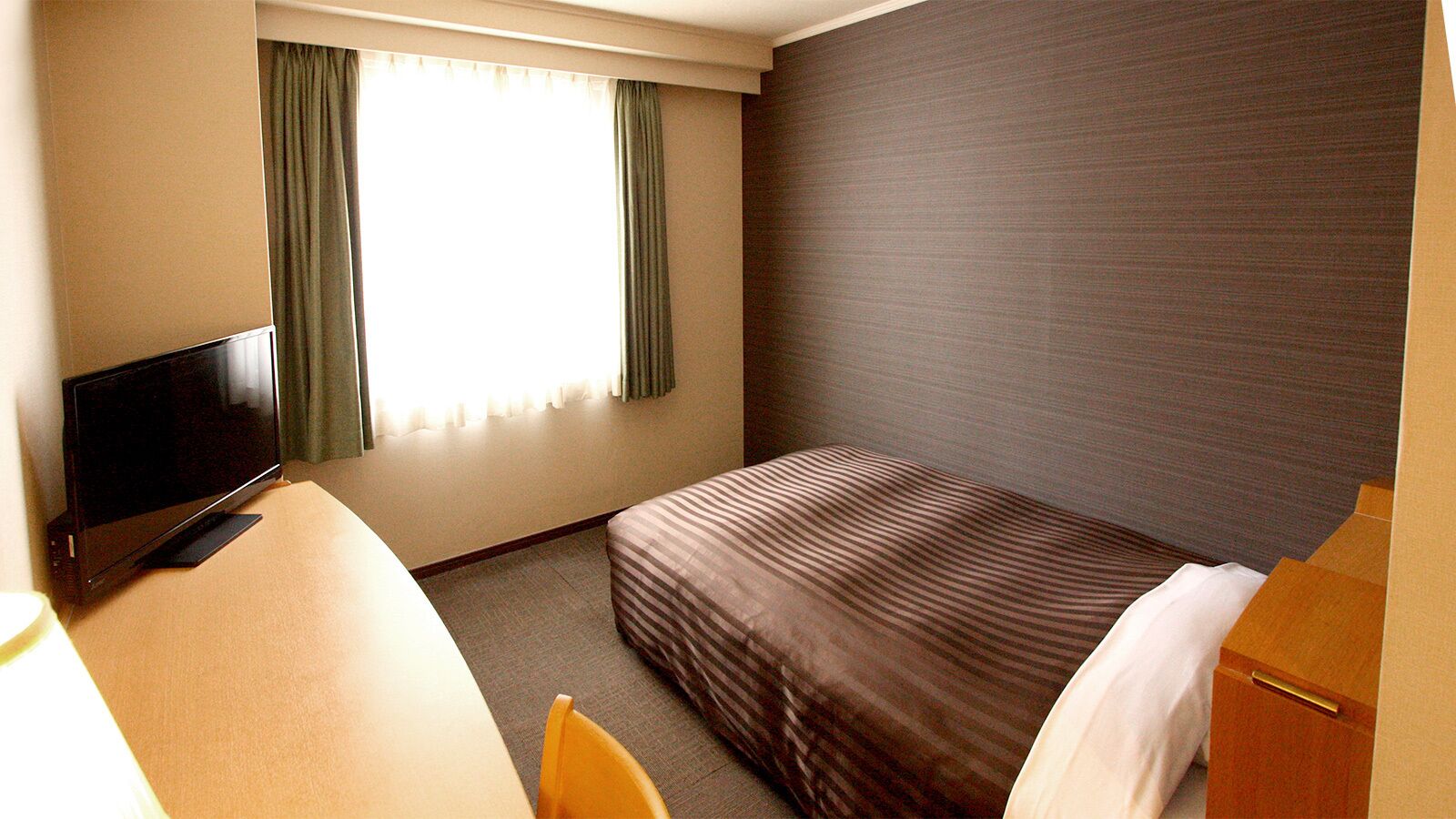 A hotel room with a bed, a desk with a TV, a chair, and a window with curtains.