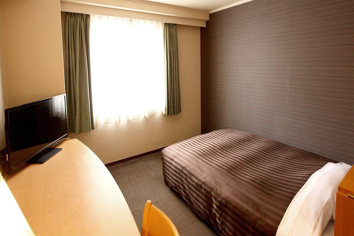 A hotel room with a bed, a desk with a TV, a chair, and a window with curtains.