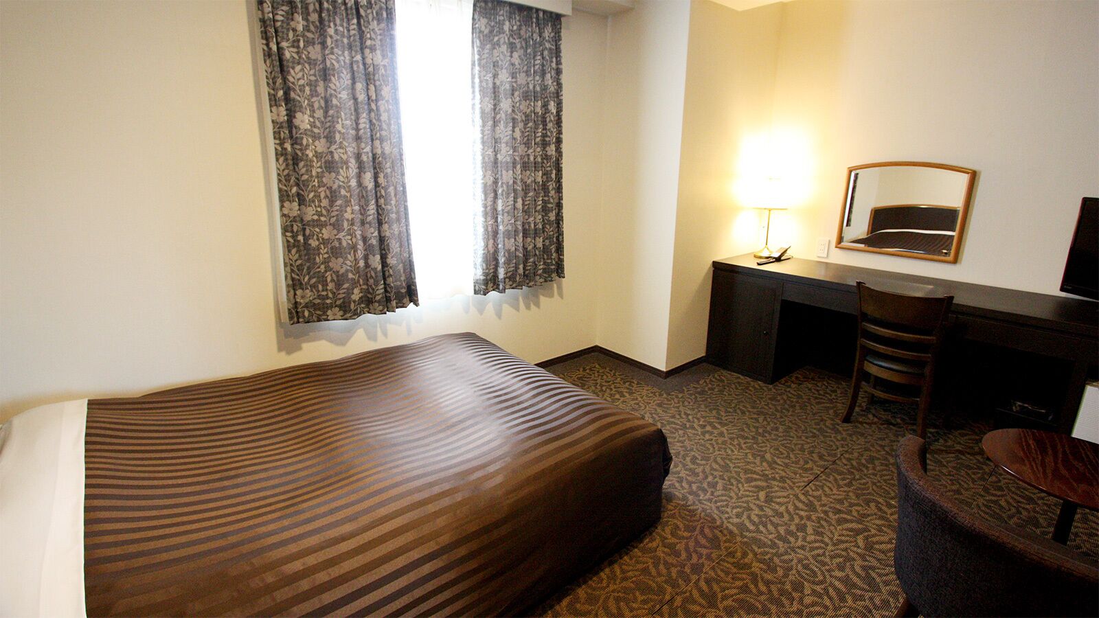 A hotel room with a bed, a desk, a chair, a lamp, and a mirror.