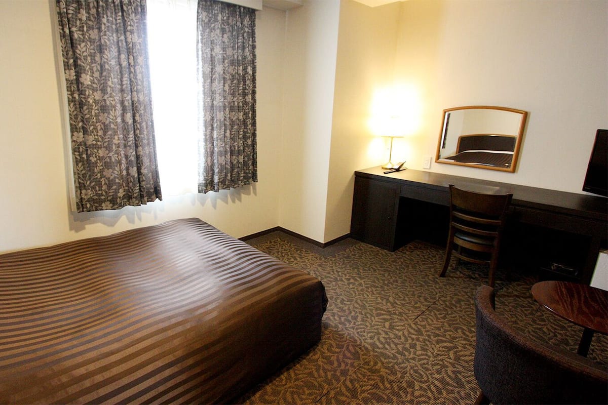 A hotel room with a bed, a desk, a chair, a lamp, and a mirror.