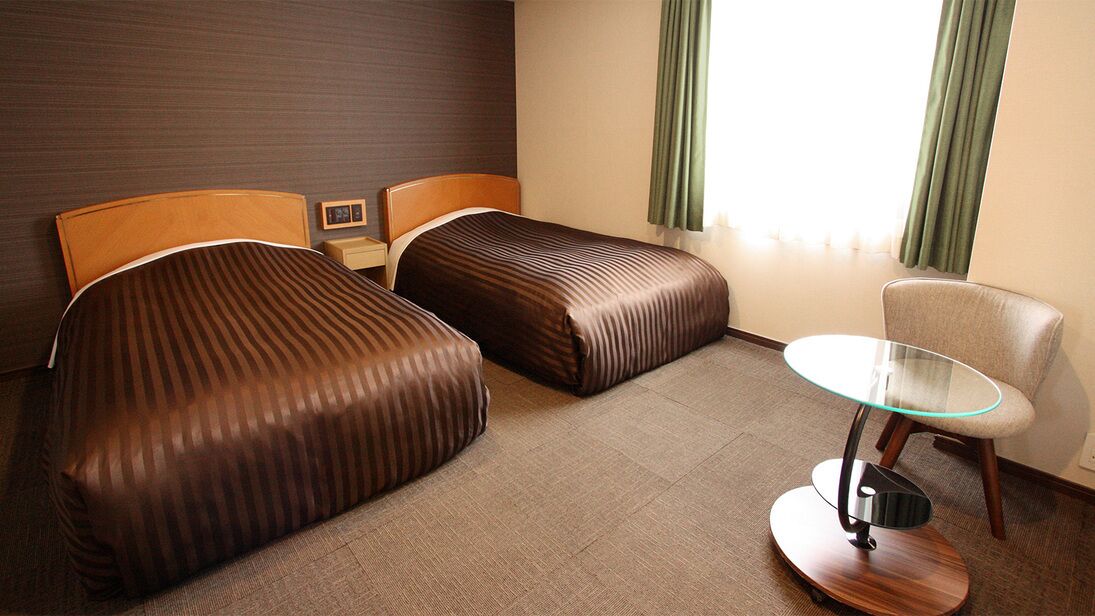 A hotel room with two beds, a small table, a chair, and a window with curtains.
