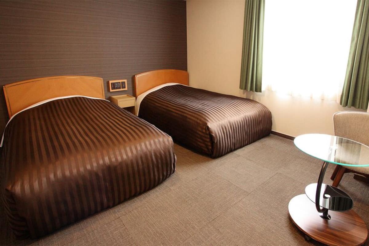 A hotel room with two beds, a small table, a chair, and a window with curtains.