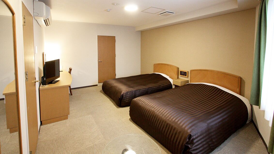 A hotel room with two beds, a desk with a computer, a door, and a window with curtains.