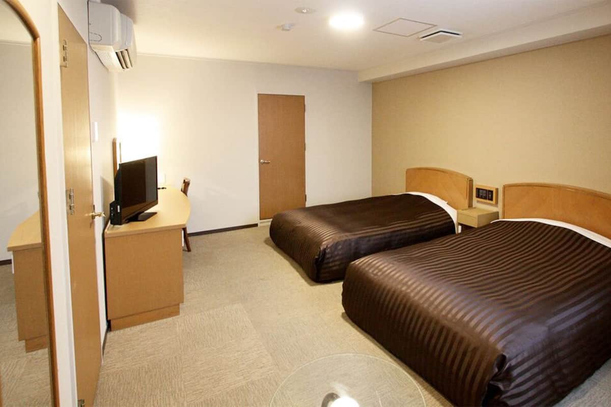 A hotel room with two beds, a desk with a computer, a door, and a window with curtains.
