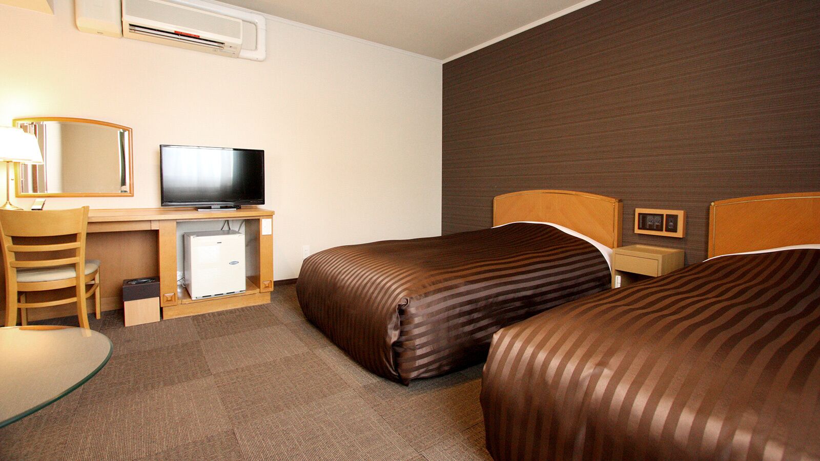 A hotel room with two beds, a desk with a television, a chair, a small table, and a bedside table with lamps.