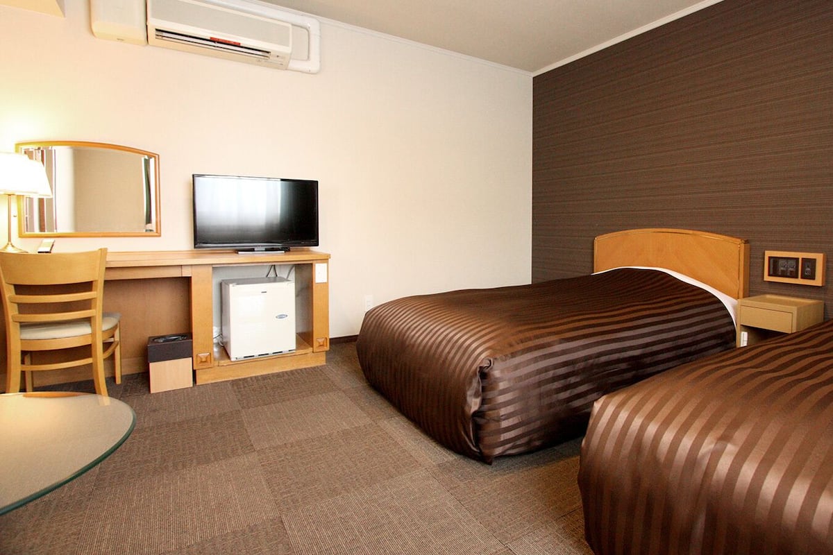 A hotel room with two beds, a desk with a television, a chair, a small table, and a bedside table with lamps.
