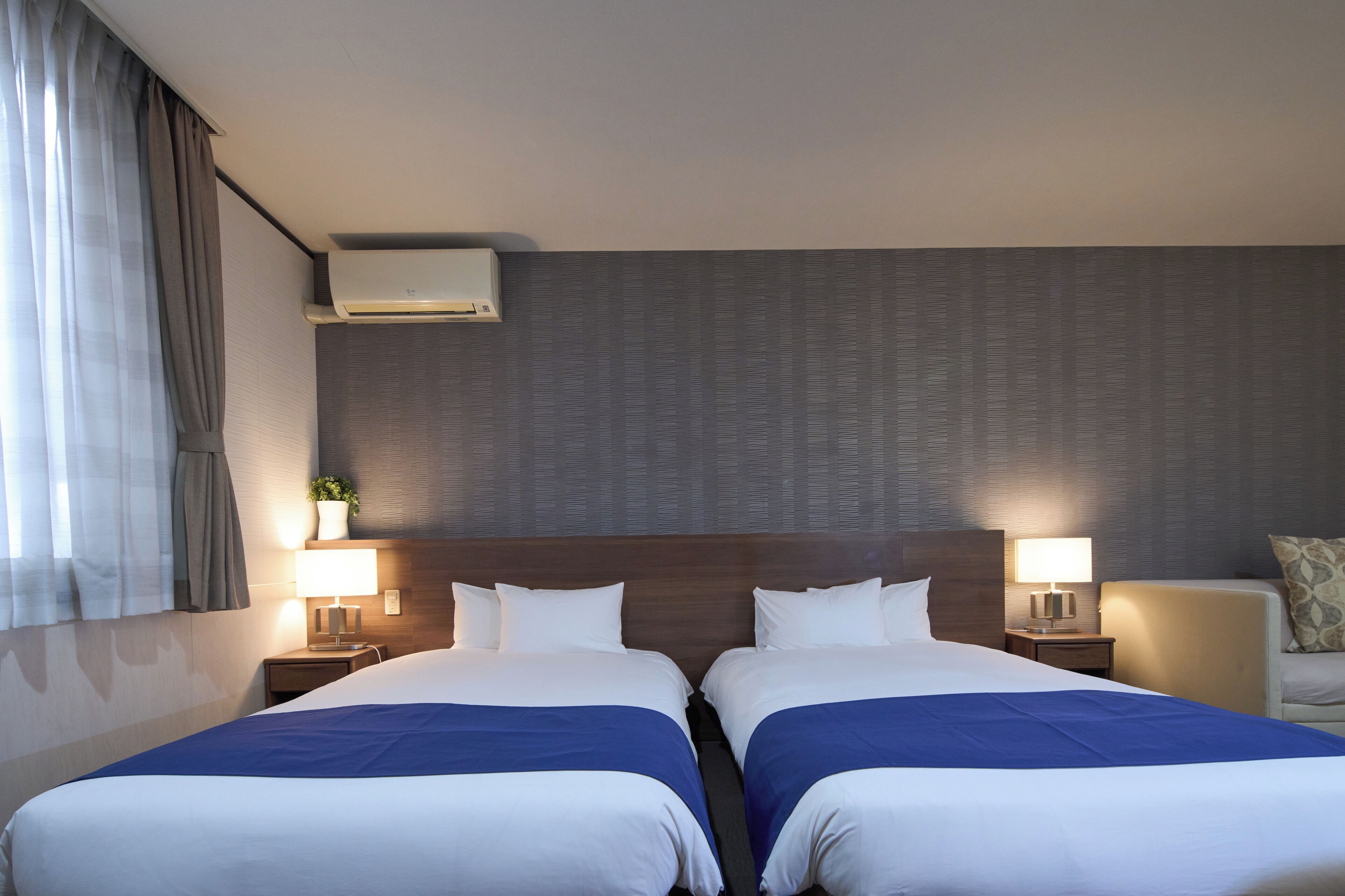 A hotel room with two beds, a large window with curtains, an air conditioning unit, and bedside lamps.