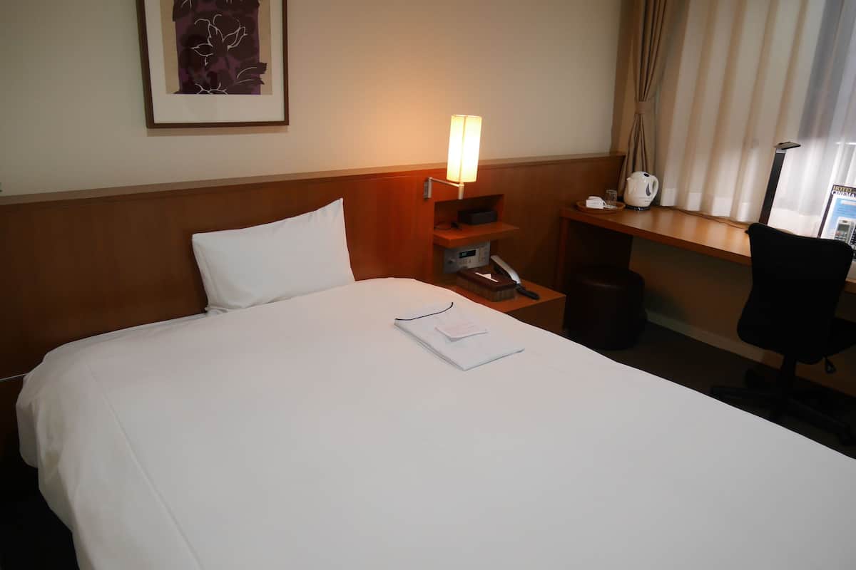 A hotel room with a neatly made bed, a bedside lamp, a small table with a kettle, and a chair.