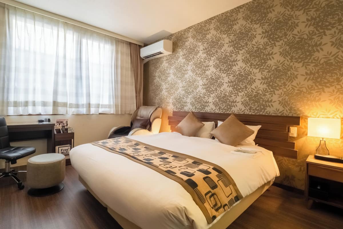 A hotel room with a bed, a desk, a chair, a massage chair, and a lamp.