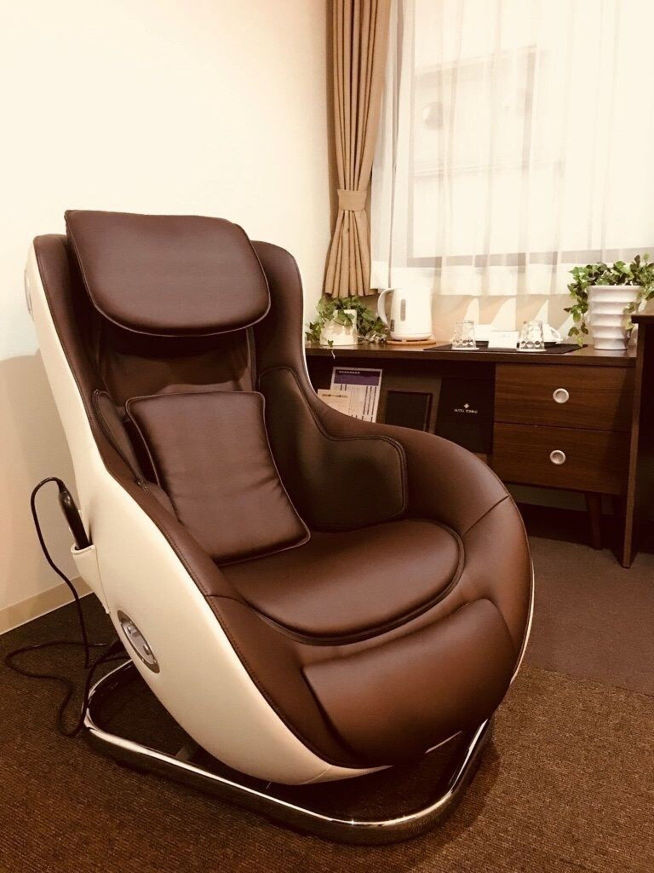 Ein Massagesessel in einem Raum mit Schreibtisch, Blumentopf und einem fenster mit Vorhängen.