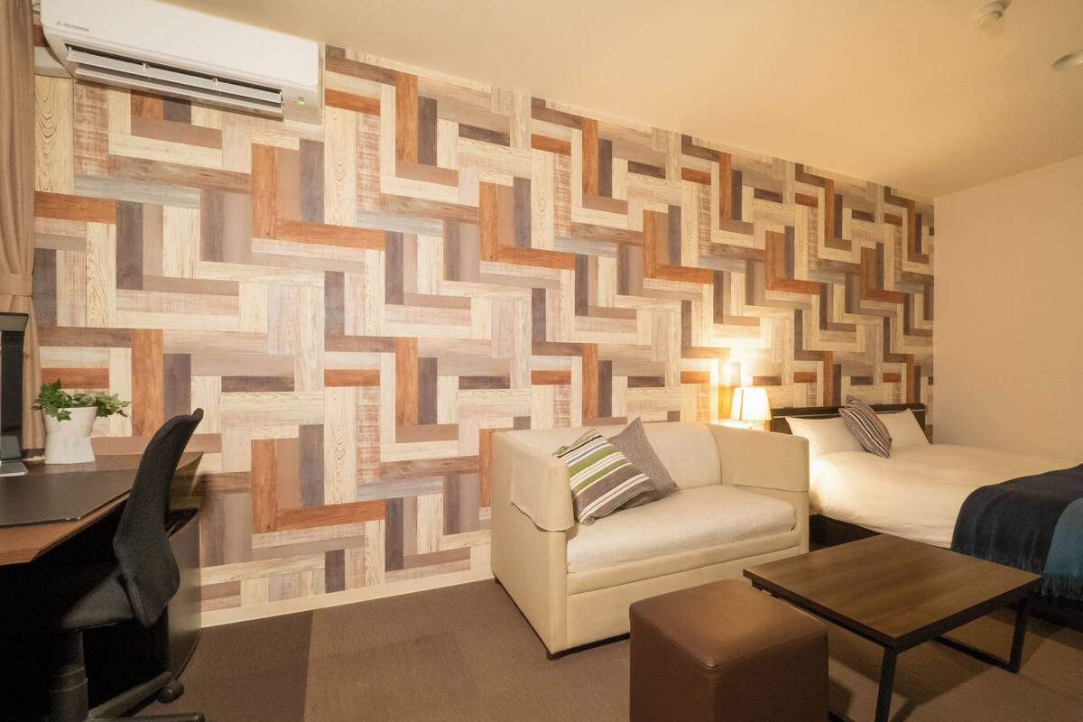 A hotel room with a modern geometric-patterned wall, a sofa, a bed, a small table, and a desk with a chair.