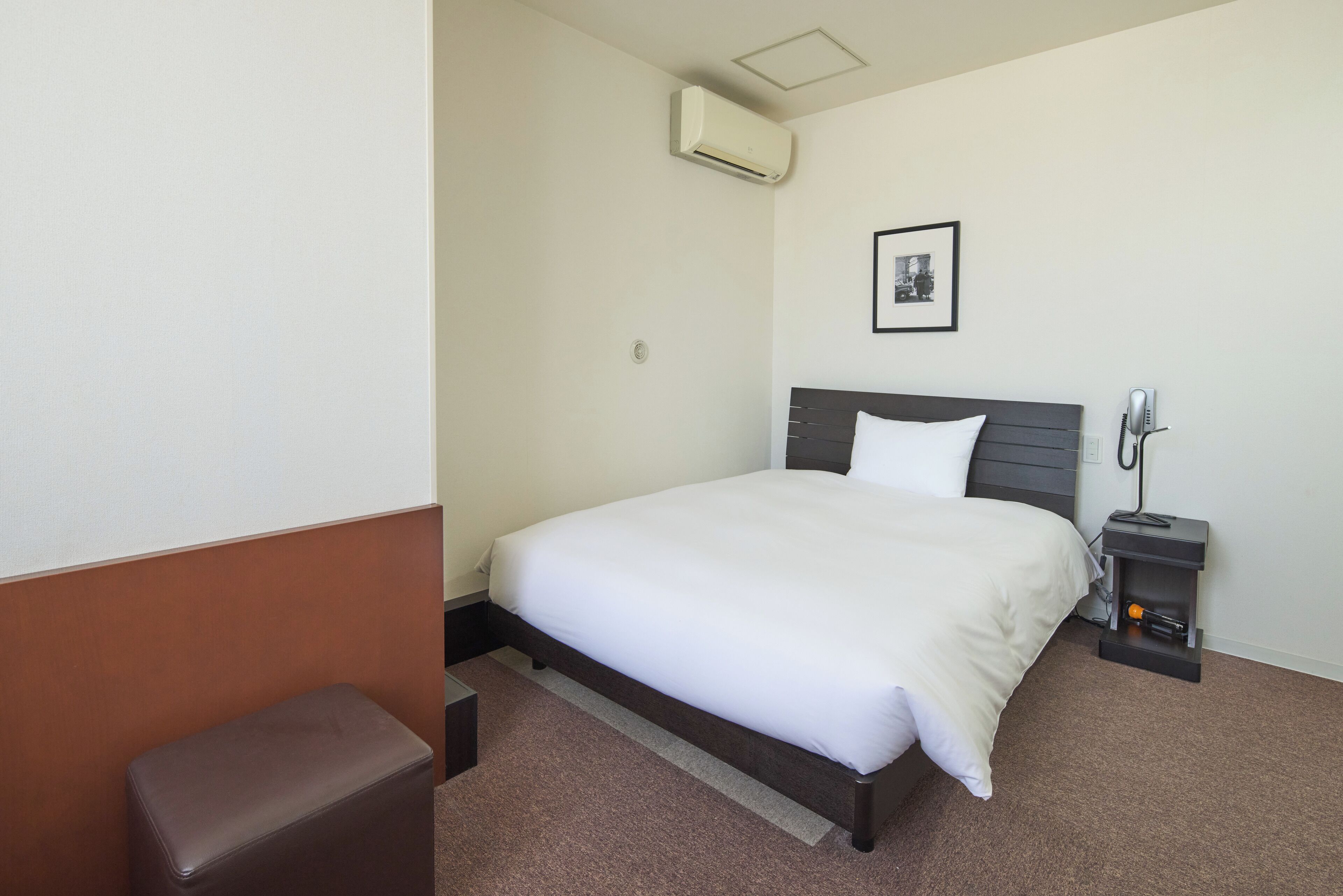 A hotel room with a bed, a bedside table with a lamp, a small stool, and an air conditioning unit.