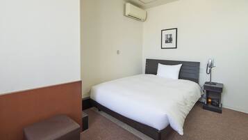 A hotel room with a bed, a bedside table with a lamp, a small stool, and an air conditioning unit.