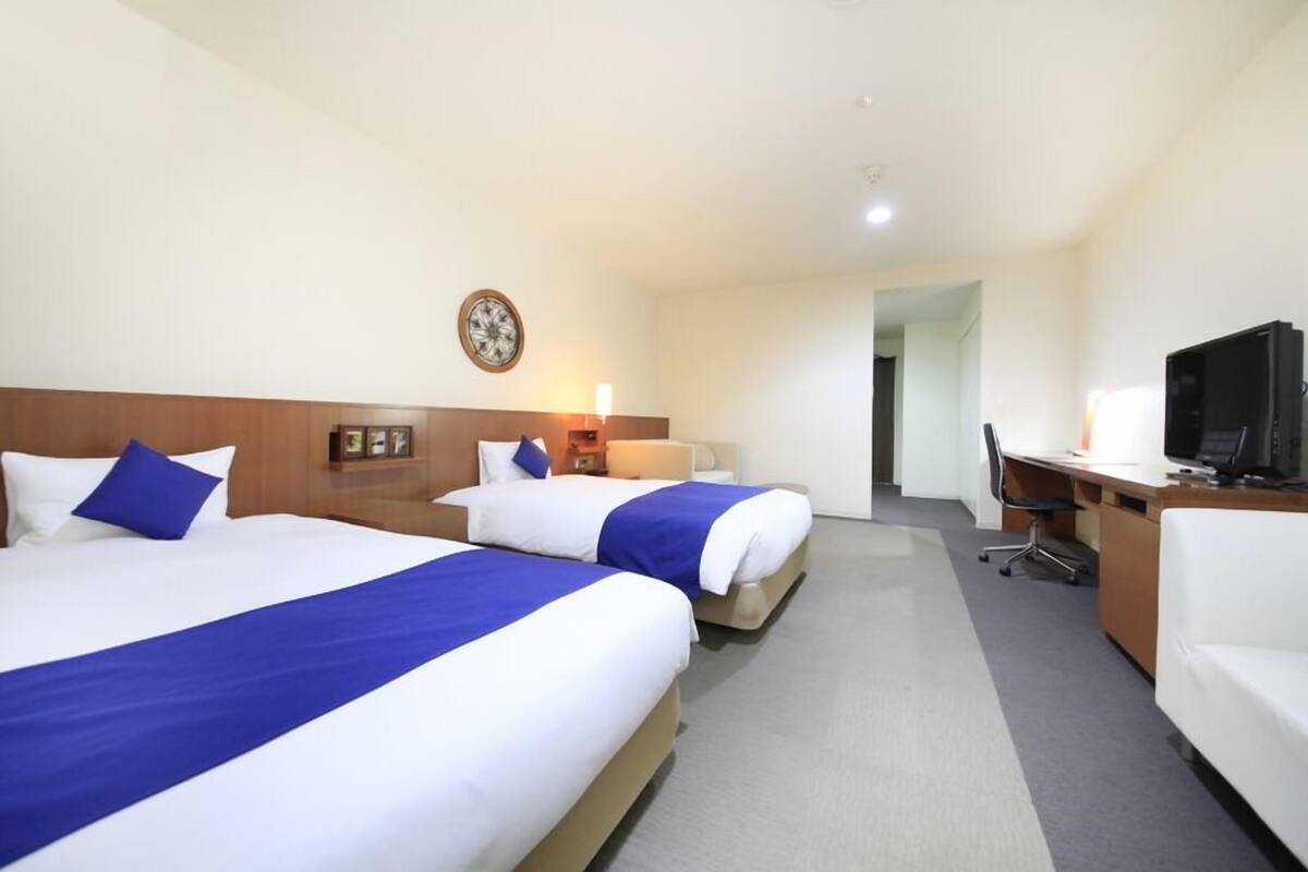 A hotel room with two beds, a desk, a chair, a television, and a clock on the wall.