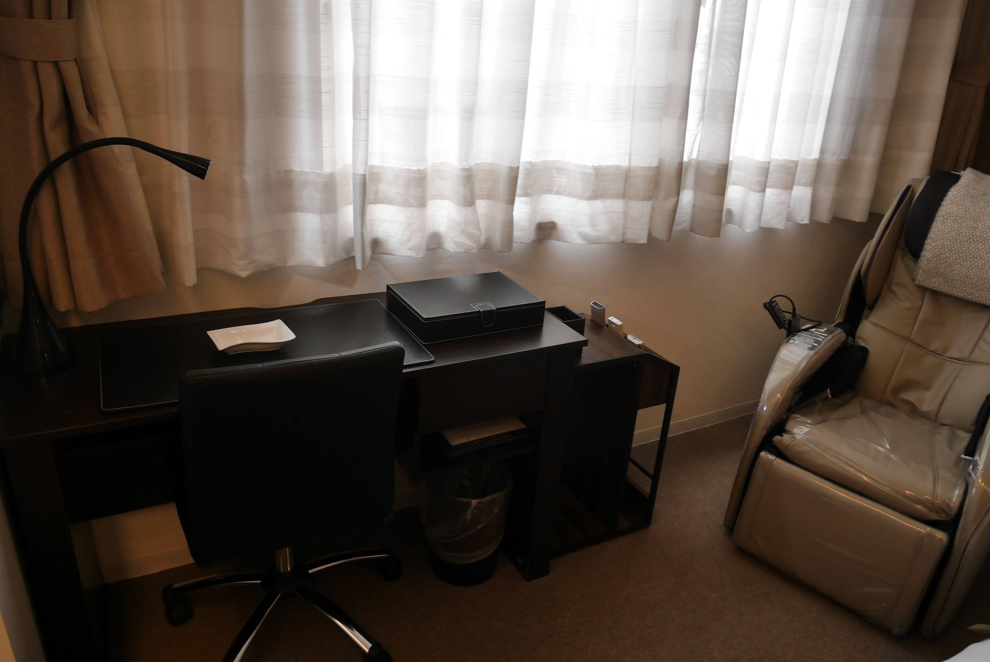 A hotel room with a desk, chair, massage chair, and a trash bin.