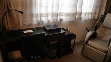 A hotel room with a desk, chair, massage chair, and a trash bin.