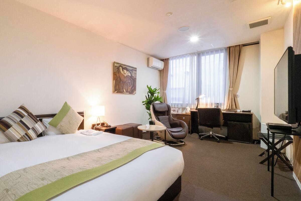 A hotel room with a large bed, a desk, a chair, a TV, and a window with curtains.