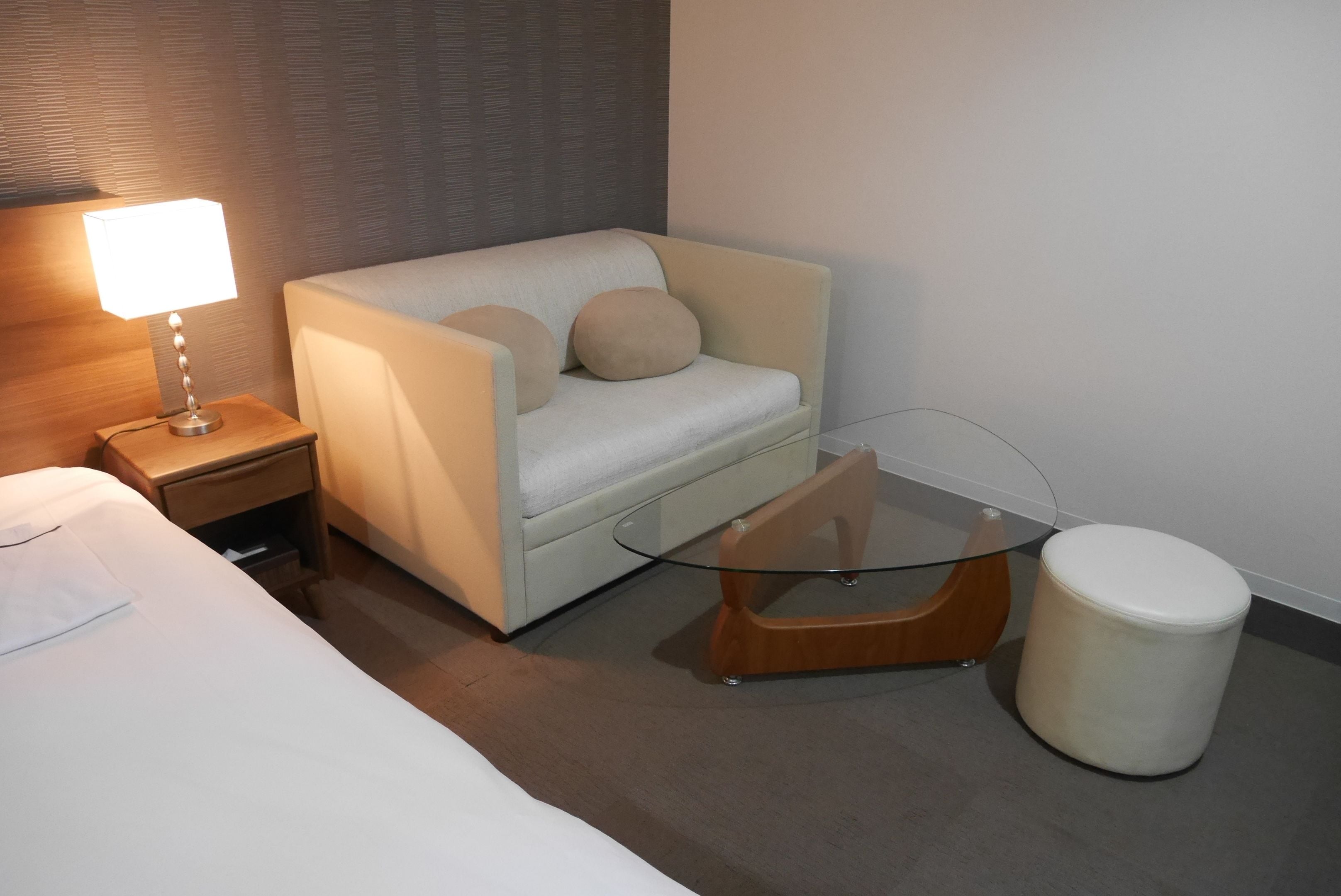 A modern hotel room with a bed, a sofa, a glass coffee table, a wooden side table, and a white trash bin.