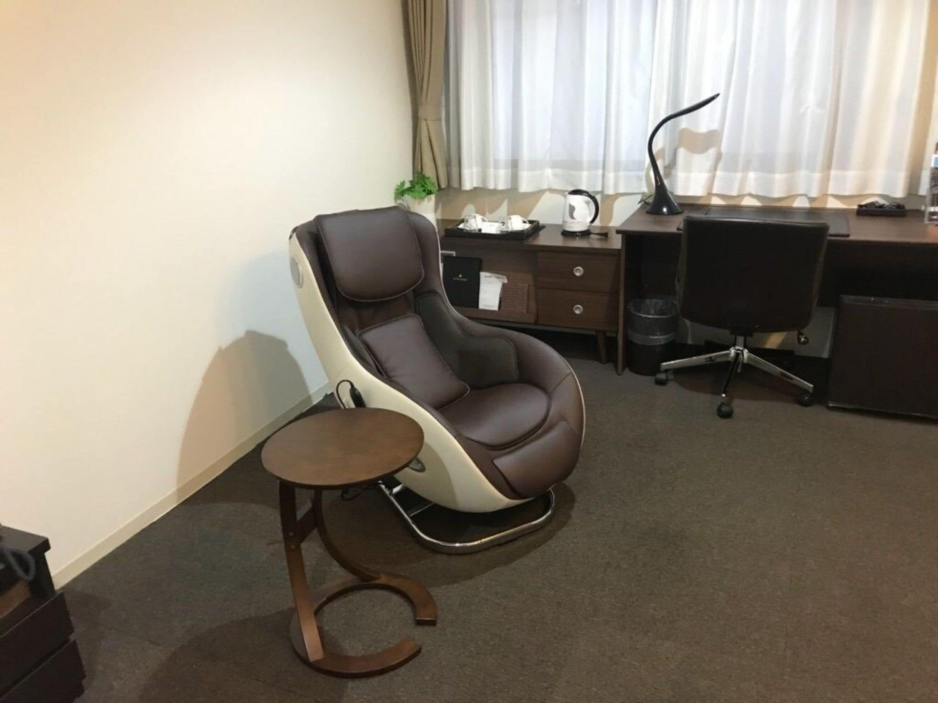 A modern office chair with a footrest, a desk with a lamp, and a trash can.