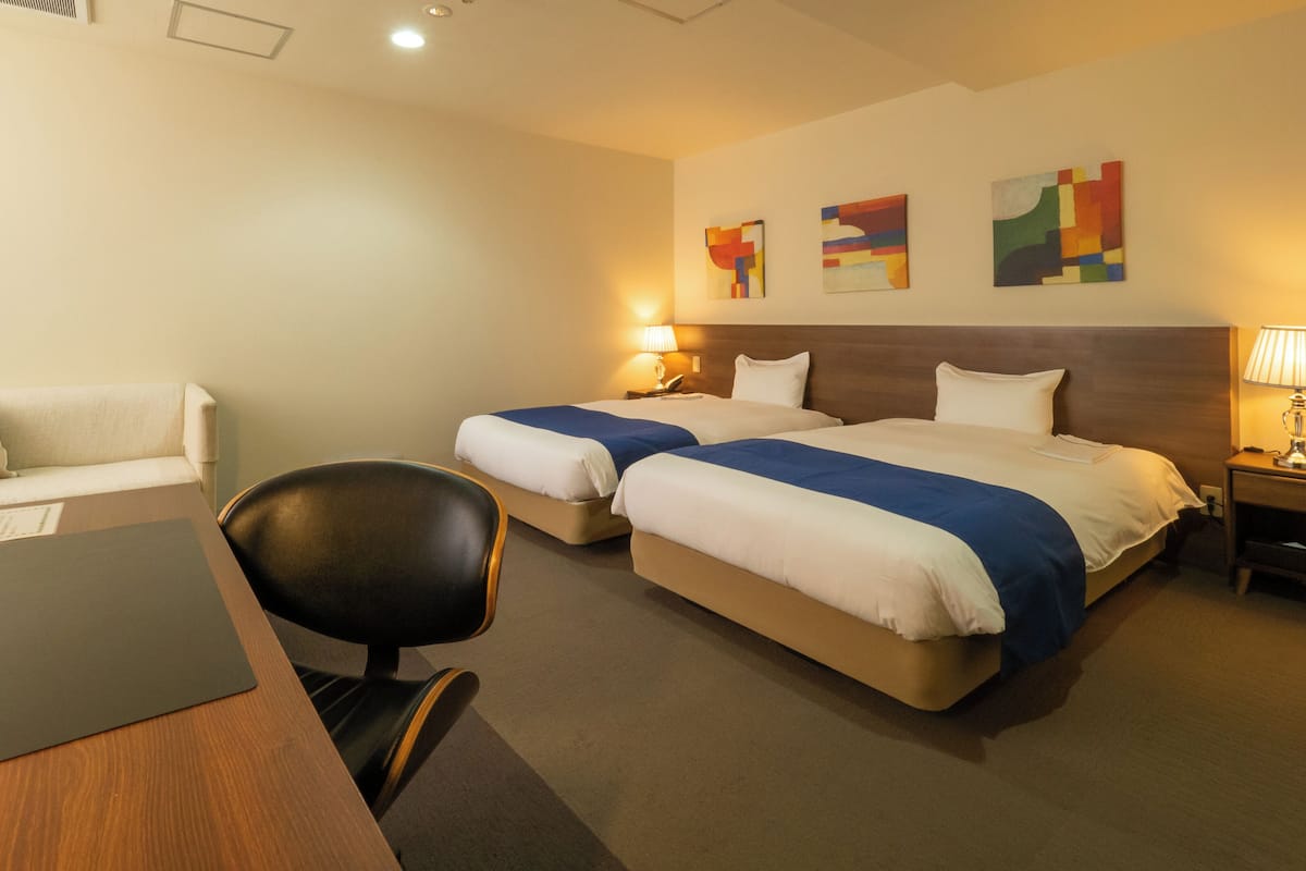 A hotel room with two beds, a desk, a chair, and a nightstand with a lamp.