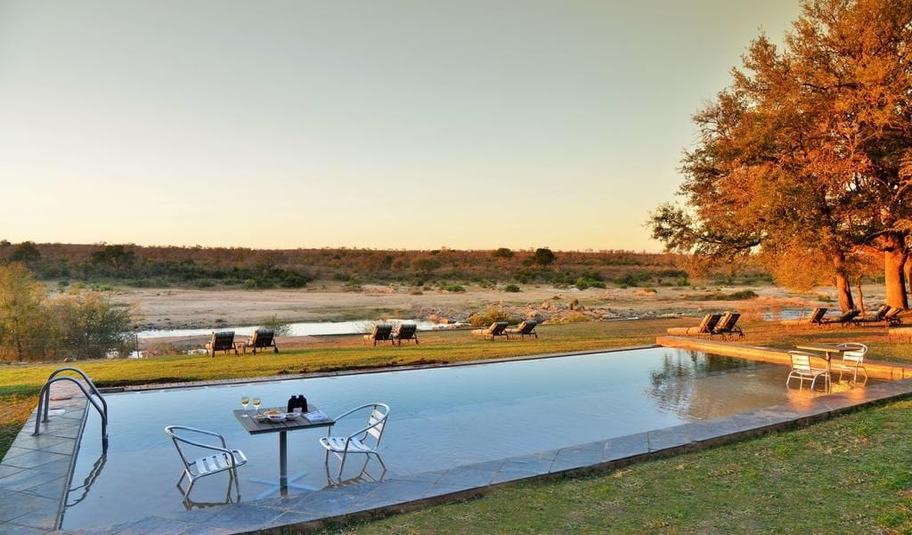 A swimming pool with lounge chairs and a table, surrounded by grass and trees.