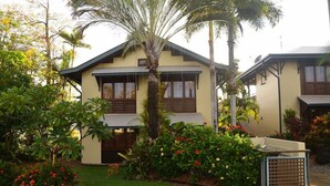 A two-story building with a yellow facade, wooden shutters, and a small garden with palm trees and flowering plants. - The Wongalinga Apartments (Wongaling Beach)