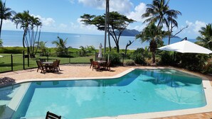 A swimming pool with a view of the ocean, surrounded by palm trees and a fenced patio area with outdoor furniture. - The Wongalinga Apartments (Wongaling Beach)