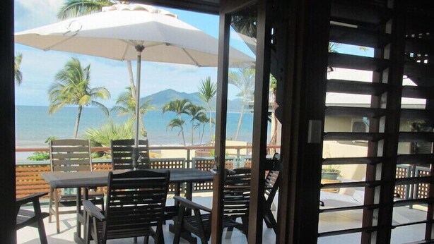 An outdoor seating area with tables and chairs, a large umbrella, and a view of the beach and mountains. - The Wongalinga Apartments (Wongaling Beach)