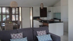 A modern living room with a grey sofa, patterned cushions, a ceiling fan, and a kitchen area with white cabinets and a black countertop. - The Wongalinga Apartments (Wongaling Beach)