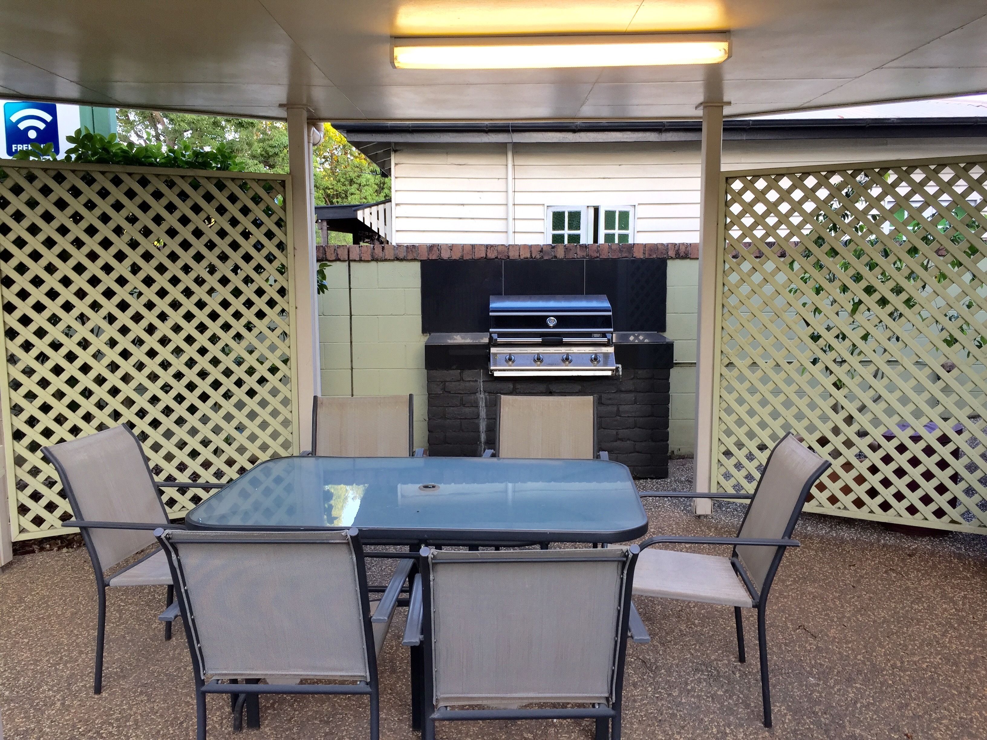 An outdoor patio with a glass table and metal chairs, a barbecue grill, and a view of a building in the background.