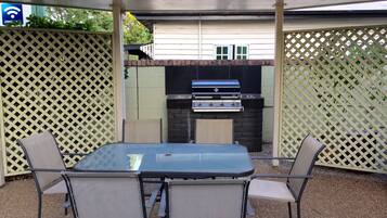 An outdoor patio with a glass table and metal chairs, a barbecue grill, and a view of a building in the background.