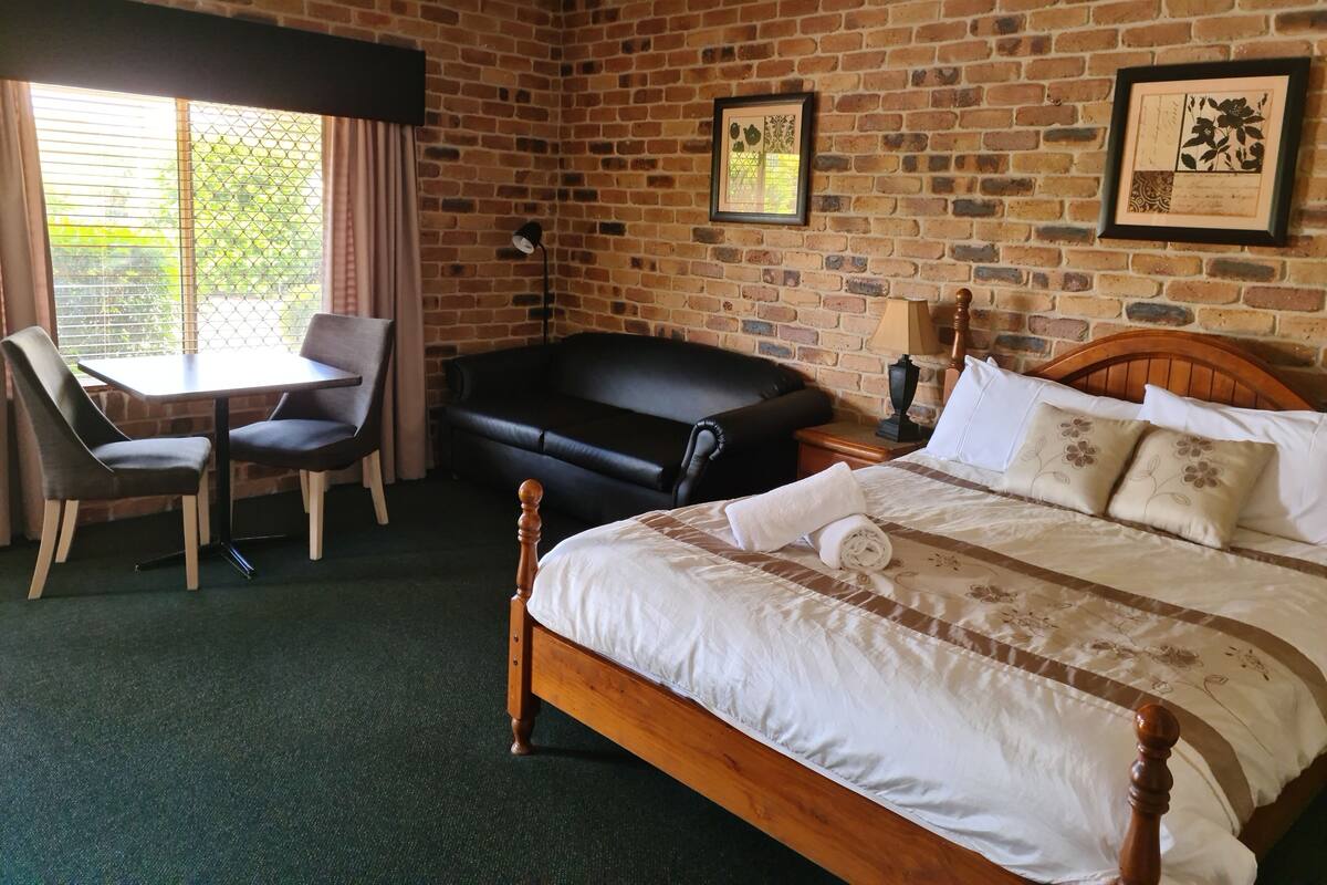 A hotel room with a brick wall, a bed with white bedding, a black leather sofa, a wooden table with two chairs, and a window with curtains.